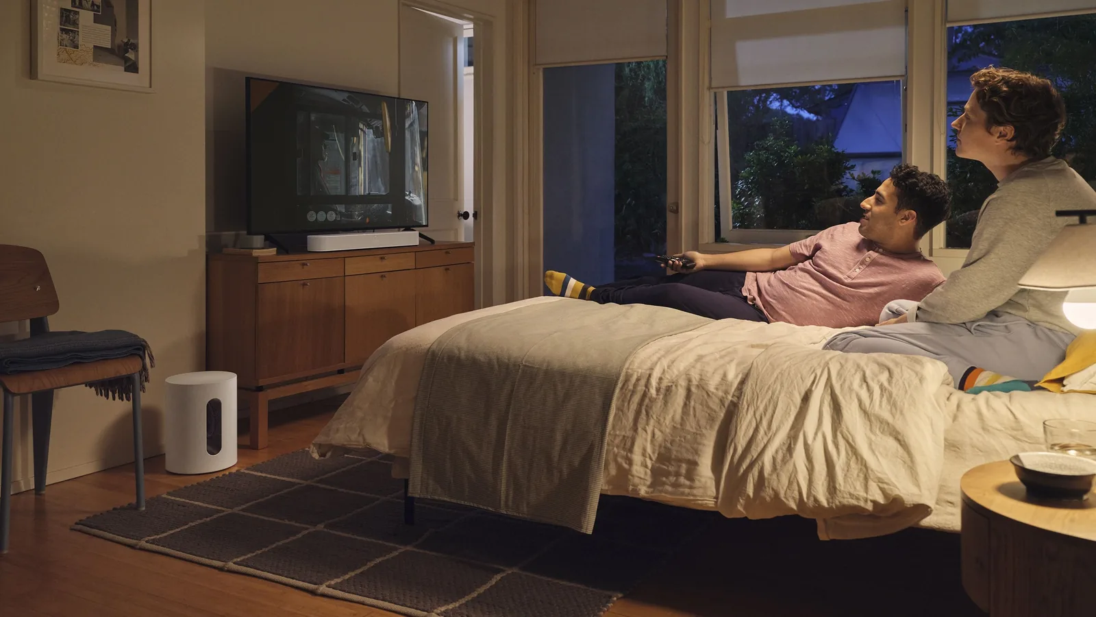Sonos Sub Mini on the floor of a bedroom beside a TV stand while two people watch a film